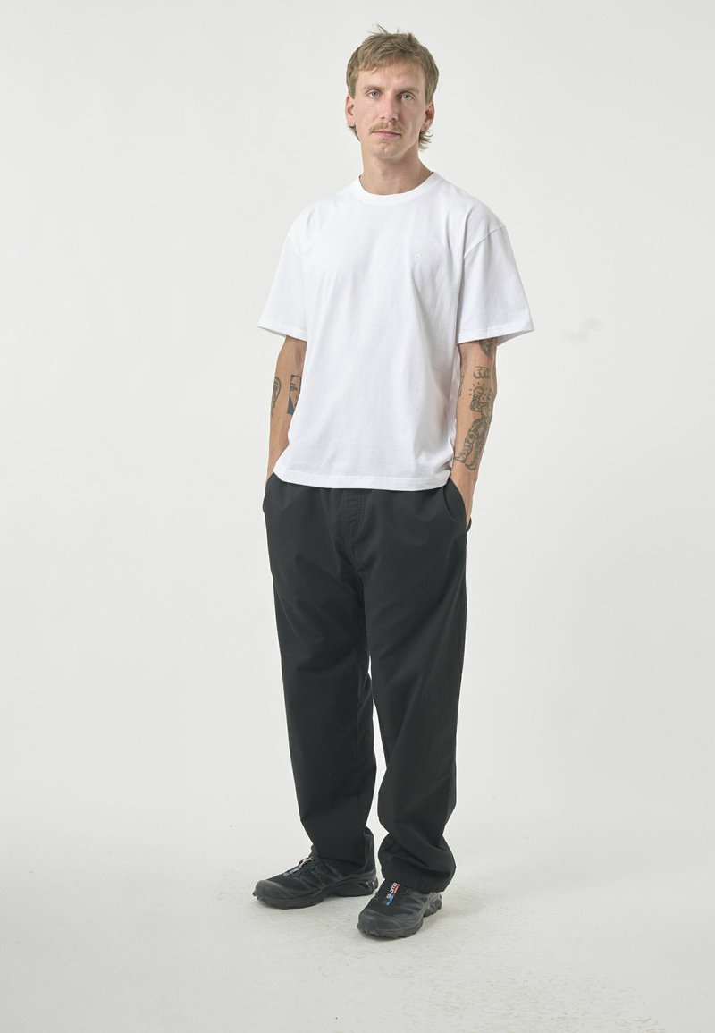 Weißes T-Shirt mit kurzen Ärmeln aus Baumwolle, kombiniert mit schwarzen, lässig sitzenden Hosen. Benutzer trägt schwarze Sportschuhe und steht mit den Händen in den Taschen.