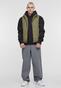 Grüne gesteppte Weste über einem schwarzen Kapuzenpullover, kombiniert mit lockeren grauen Jeans und weißen Sneakern. Glatte Textur, keine sichtbaren Muster.