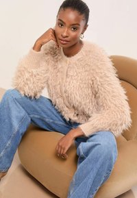 Bolyhos bézs kardigán kerek nyakkivágással és gombos záródással, világoskék, bő szárú farmernadrággal párosítva, egy világosbarna, kerek székben ülve.