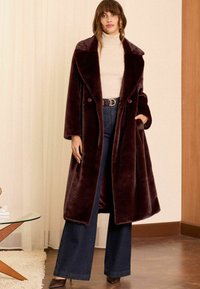 Cappotto in pelliccia sintetica bordeaux con colletto oversize, bottoni doppi e maniche lunghe. Indossato sopra un maglione a collo alto chiaro e jeans a gamba larga.