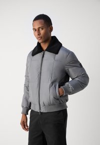 Veste rembourrée grise avec un col en fourrure noire, fermeture éclair à l'avant, poignets élastiques et poches latérales. Présente une silhouette carrée et un design matelassé.