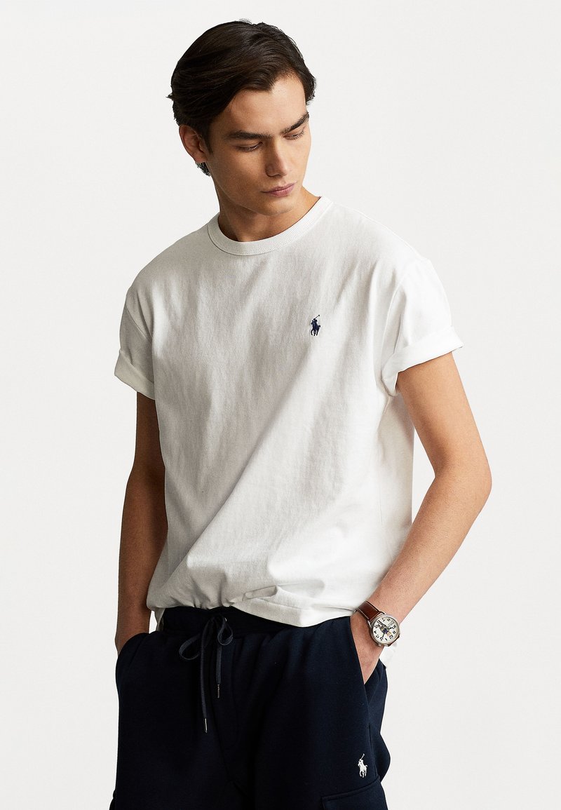 T-shirt bianco in cotone con collo a giro e maniche arrotolate, caratterizzato da un piccolo logo blu navy sul petto. Abbinato a pantaloni navy e un orologio.