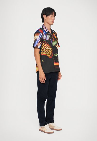 Camicetta a maniche corte con una stampa astratta vibrante e multicolore; sfondo nero, design con bottoni, abbinata a pantaloni scuri e sneakers chiare.