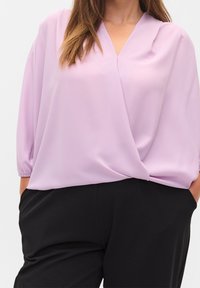 Blusa lavanda con scollo a V, maniche a tre quarti e polsini elasticizzati. Tessuto morbido che cade delicatamente, abbinato a pantaloni neri.