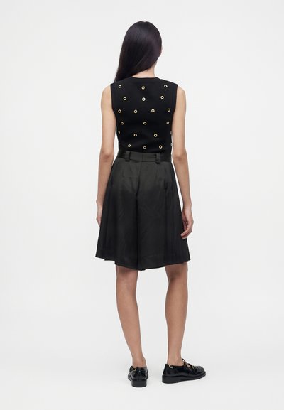Femme aux longs cheveux foncés portant un haut noir sans manches avec des œillets dorés et un short noir taille haute à jambes larges, debout de dos.