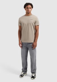 Beige T-shirt met korte mouwen en een ronde halslijn, subtiele logo-detail, in combinatie met grijze recht gesneden jeans en zwart-witte sneakers.