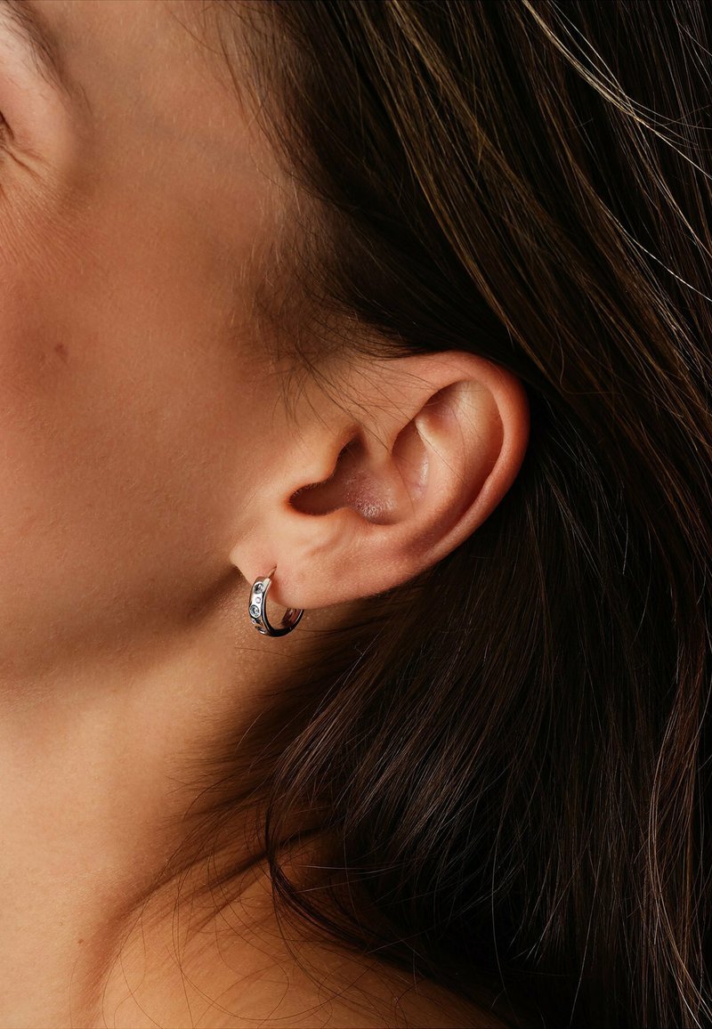 Silberner Creolenohrring mit drei kleinen klaren Steinen, glatter Oberfläche und leicht gekrümmtem Design. Getragen am Ohr einer Person mit dunklem Haar.