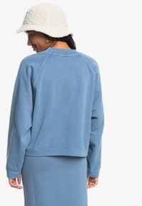 Felpa blu con colletto a girocollo e maniche raglan, realizzata in morbido cotone. Presenta polsini e orlo a coste; indossata con una gonna coordinata.