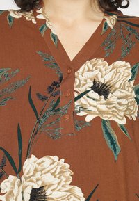 Brązowa bluzka w kwiaty z dużymi kremowymi kwiatami i zielonymi liśćmi, z dekoltem w kształcie litery V oraz czterema brązowymi guzikami z przodu. Gładka faktura materiału.