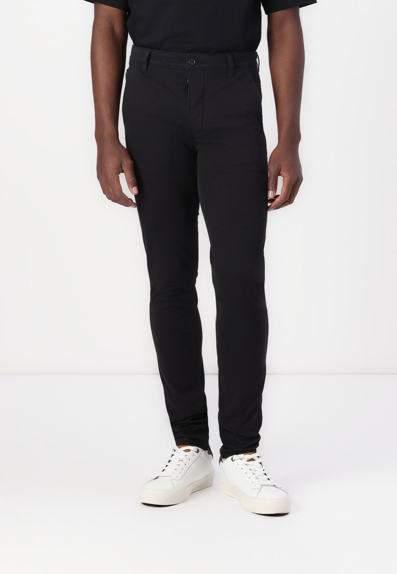 Homme portant un pantalon noir slim et des baskets blanches, debout devant un fond blanc.