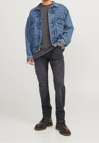 Blauwe denimbomberjack met knopen en opgestikte zakken, gecombineerd met een donkergrijze t-shirt en zwarte jeans, aangevuld met een paar bruine laarzen.