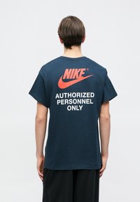 Ναυτικό βαμβακερό μπλουζάκι με κοντά μανίκια, που διαθέτει ένα μεγάλο κόκκινο λογότυπο Nike και λευκό κείμενο "ΜΟΝΟ ΓΙΑ ΕΞΟΥΣΙΟΔΟΤΗΜΕΝΑ ΑΤΟΜΑ" στην πλάτη.