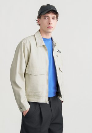 Beige lærredsjakke med krave, to brystlommer og et sort logo-mærke. Båret over en blå skjorte og kombineret med sorte bukser og kasket.