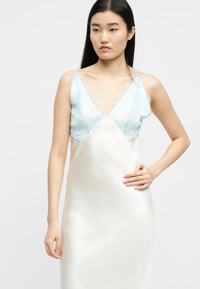 Νεαρή γυναίκα με μακριά μαύρα μαλλιά που φοράει ένα λευκό φόρεμα με λεπτές τιράντες και ανοιχτά μπλε δαντελένια ντεκολτέ, στέκεται με το ένα χέρι στη μέση.