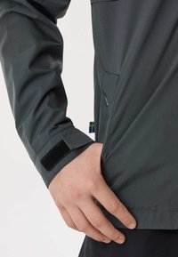 Roka delno v stranskem žepu temno sive jakne z manšetnim trakom na ježka in majhno modro-rumeno tkanino oznako.