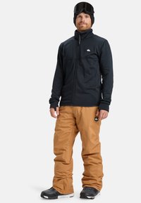Čierna flisová bunda na zips, hnedé snežné nohavice a čierne snowboardové topánky. Obsahuje lyžiarske okuliare a čiapku, pričom prezentuje teplý dizajn.