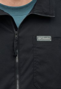 Κοντινό πλάνο ενός μαύρου τζάκετ Columbia με φερμουάρ που φοριέται πάνω από μια τιρκουάζ μπλούζα, δείχνοντας το φερμουάρ και μια τσέπη στο στήθος με την ετικέτα της μάρκας.