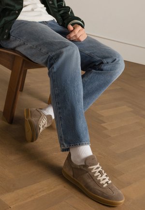 Baskets en suède beige avec des lacets blancs et des semelles en gomme, associées à un jean en denim bleu clair et des chaussettes blanches, assises sur une chaise en bois.