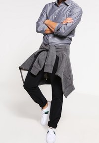 Šedá košile na knoflíky s malým logem, černé kalhoty a bílé tenisky se zelenými akcenty. Šedá mikina uvázaná v pase.