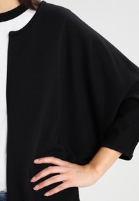 Svart oversized topp i mjukt tyg, med vid halsringning, korta ärmar och en lös passform. Sömdetalj längs den främre delen.