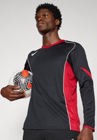 Cămașă sportivă neagră cu mânecă lungă, cu accente din plasă roșie și cusături albe, purtată în timp ce ține un minge de fotbal cu un model, pe un fundal neutru.