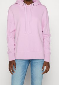 Light pink hoodie made of soft fabric, featuring a drawstring hood, ribbed cuffs, and a relaxed fit. Paired with light blue jeans.