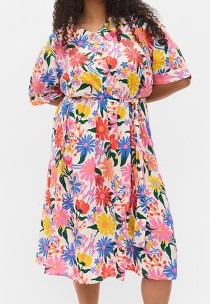 Vrouw draagt een knielange jurk met riem, korte mouwen en een meerkleurig bloemenpatroon op een lichte achtergrond.