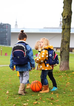 Drei Kinder mit Rucksäcken im Haifischdesign: einer in Marineblau mit roten Akzenten, einer mit gelbem gepunktetem Mantel und ein orangefarbener Basketball auf dem Gras.