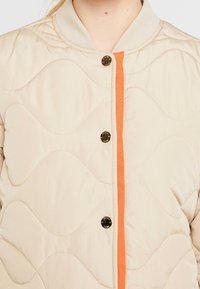 Prošívaná béžová bunda s hladkým povrchem, která má oranžový vertikální pruh, šedý ribbed límec a černé patentky.