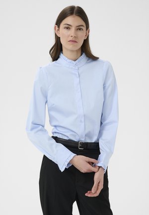 Frau trägt eine hellblaue Bluse mit Knöpfen, Rüschen am Kragen und an den Manschetten, schwarzen Gürtel und schwarze Hose und richtet ihre linke Ärmel.