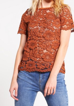 Žena v rezavě hnědém květinovém krajkovém topu s krátkými rukávy a modrých džínách, stojící před bílým pozadím.