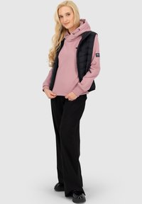 Donna bionda che indossa una felpa mauve, un gilet imbottito nero, pantaloni larghi neri e scarpe nere, in piedi con le mani che aggiustano l'orlo della felpa.