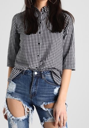 Žena oblečená v černo-bílé kostkované košili s knoflíky a tříčtvrtečními rukávy a modrých džínách s výraznými dírami.