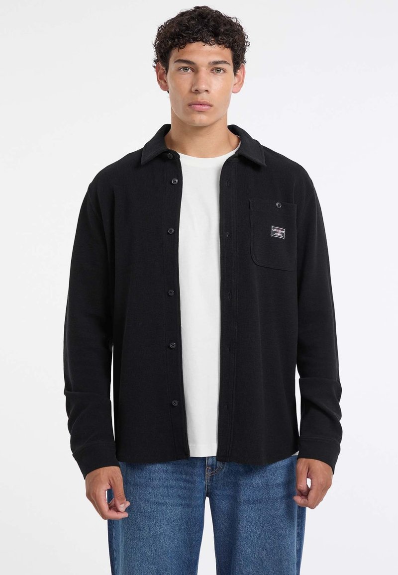 Uomo con capelli ricci che indossa una camicia nera con bottoni sopra una maglietta bianca e jeans blu, in piedi contro uno sfondo bianco.
