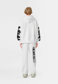 Sudadera blanca con detalles gráficos negros en las mangas, combinada con pantalones blancos que presentan un diseño impreso en el lateral. Tejido texturizado, ajuste relajado.
