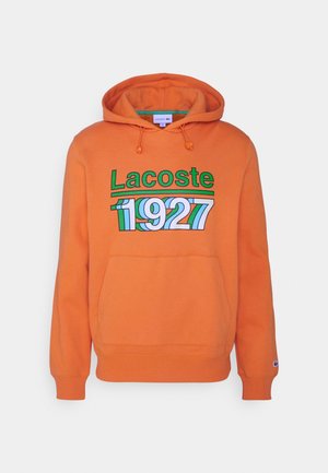 Pomarańczowa bluza z kapturem i kieszenią z przodu, z zielonym napisem "Lacoste" i kolorowym nadrukiem "1927" na piersi.