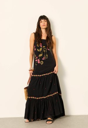 Kobieta z długimi brązowymi włosami, ubrana w czarną, bezrękawną suknię maxi z kwiatową haftem, trzyma plecioną torbę i nosi czarne sandały.