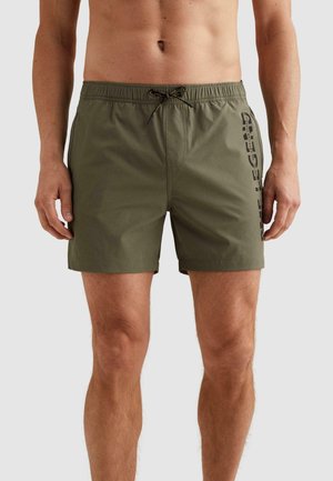 Muž nosiaci olivovo zelené plavkové šortky s čiernym sťahovacím šnúrkom a zvislým nápisom „DIESEL LEGEND“ na pravom stehne na jednoduchom podklade.