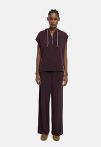 Kobieta z warkoczami, ubrana w bezrękawnik w ciemnofioletowym kolorze oraz pasujące szerokie spodnie, w brązowych mokasynach na białym tle.