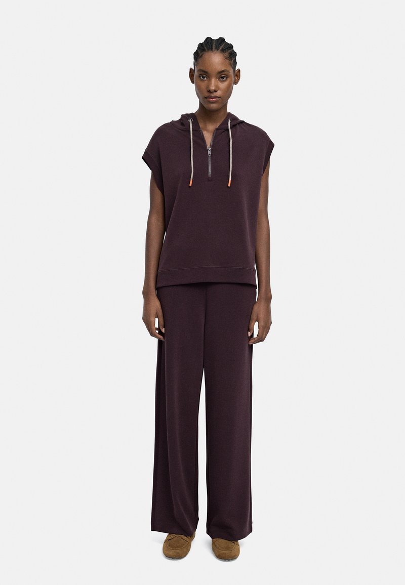 Kobieta z warkoczami, ubrana w bezrękawnik w ciemnofioletowym kolorze oraz pasujące szerokie spodnie, w brązowych mokasynach na białym tle.