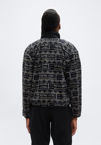 Άτομο που φοράει μαύρο και μπεζ φλις μπουφάν με σχέδιο, στέκεται με την πλάτη γυρισμένη στην κάμερα μπροστά σε απλό φωτεινό φόντο.