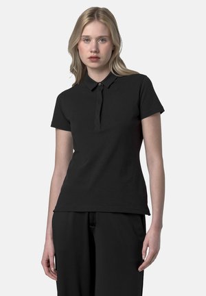 Giovane donna in piedi, indossa una maglietta polo nera aderente a maniche corte e pantaloni neri, su uno sfondo chiaro e semplice.