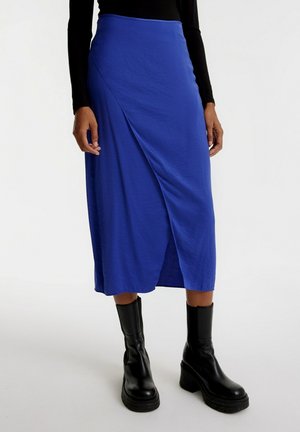 Jupe midi bleu royal avec un design croisé, fabriquée en tissu léger, dotée d'une fente sur le côté et d'une texture lisse, assortie à des bottes noires à semelle épaisse.