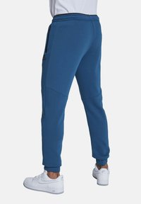 Blaue, leichte Jogginghosen mit elastischem Bund, schmal zulaufenden Beinen, seitlichen Reißverschlusstaschen und einem dezenten Logodetail nahe dem Bund.