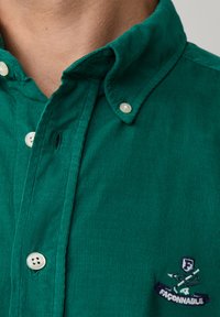 Groene corduroy overhemd met een button-down kraag, voorzien van witte knopen en een geborduurd logo aan de linkerkant bij de kraag.