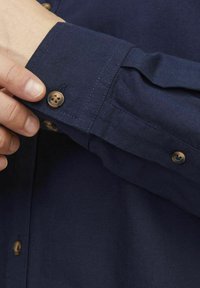 Mørkeblå skjorte med knapper og tekstiler. Har brune knapper og manchetter med stikninger.