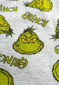 Tessuto grigio heather con illustrazioni ripetute del Grinch verde con occhi gialli e testo che recita "GRINCH" in grassetto verde.