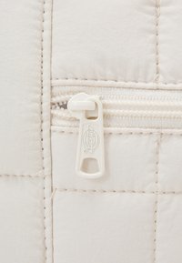 Jasno beżowa tkanina pikowana z widocznym przeszyciem, z białym zamkiem błyskawicznym i wytłoczonym logo na zawieszce zamka.