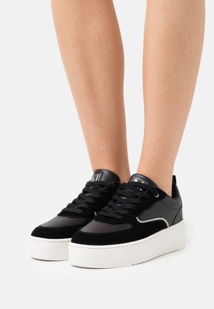 Benen met zwarte veterplateausneakers met witte dikke zolen op een effen lichte achtergrond.