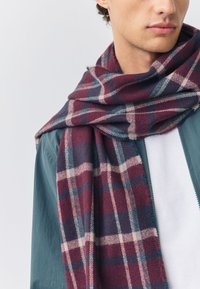 Sciarpa a quadri in rosso intenso, blu navy e crema, realizzata in tessuto morbido. Indossata in modo casual intorno al collo, presenta un design piegato con bordi frangiati.